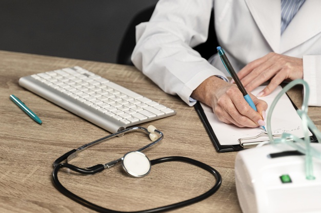 Een dokter schrijft iets op een kladblok. Op het bureau ligt een toestenbord en een stethoscoop
