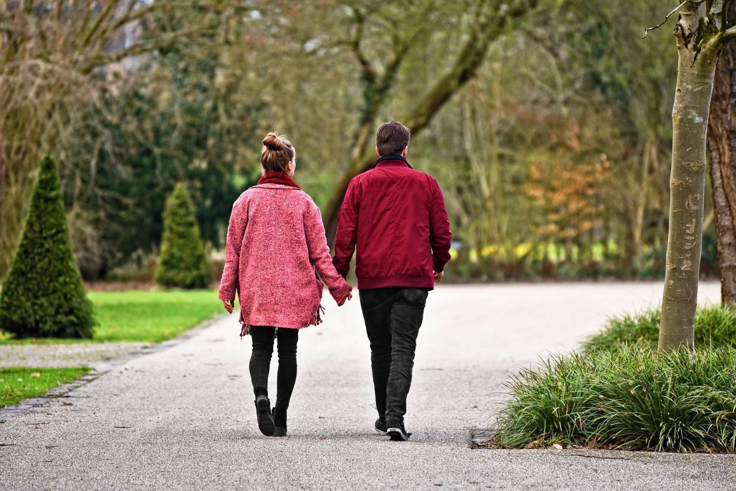 Man en vrouwen lopen hand in hand door het park. Beiden hebben een rode jas aan. De vrouw heeft haar haar in een knotje.